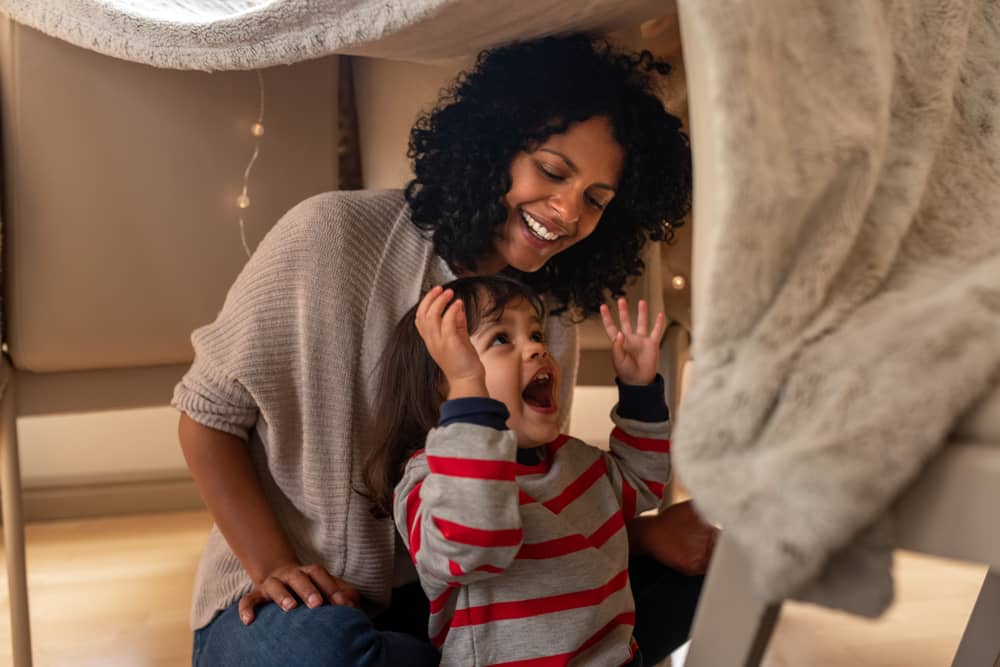 Cute little girl laughing while sitting in a blanket fort with her mother on their living room floor doing a winter activities for kids